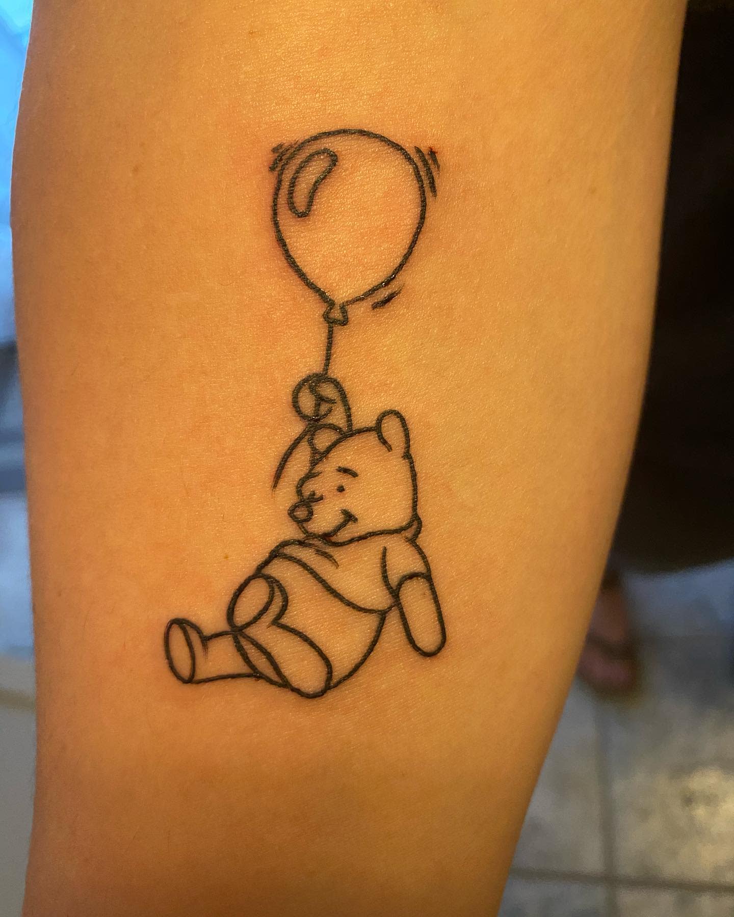 Simple outline tattoo of Winnie the Pooh floating with a balloon on a person's arm